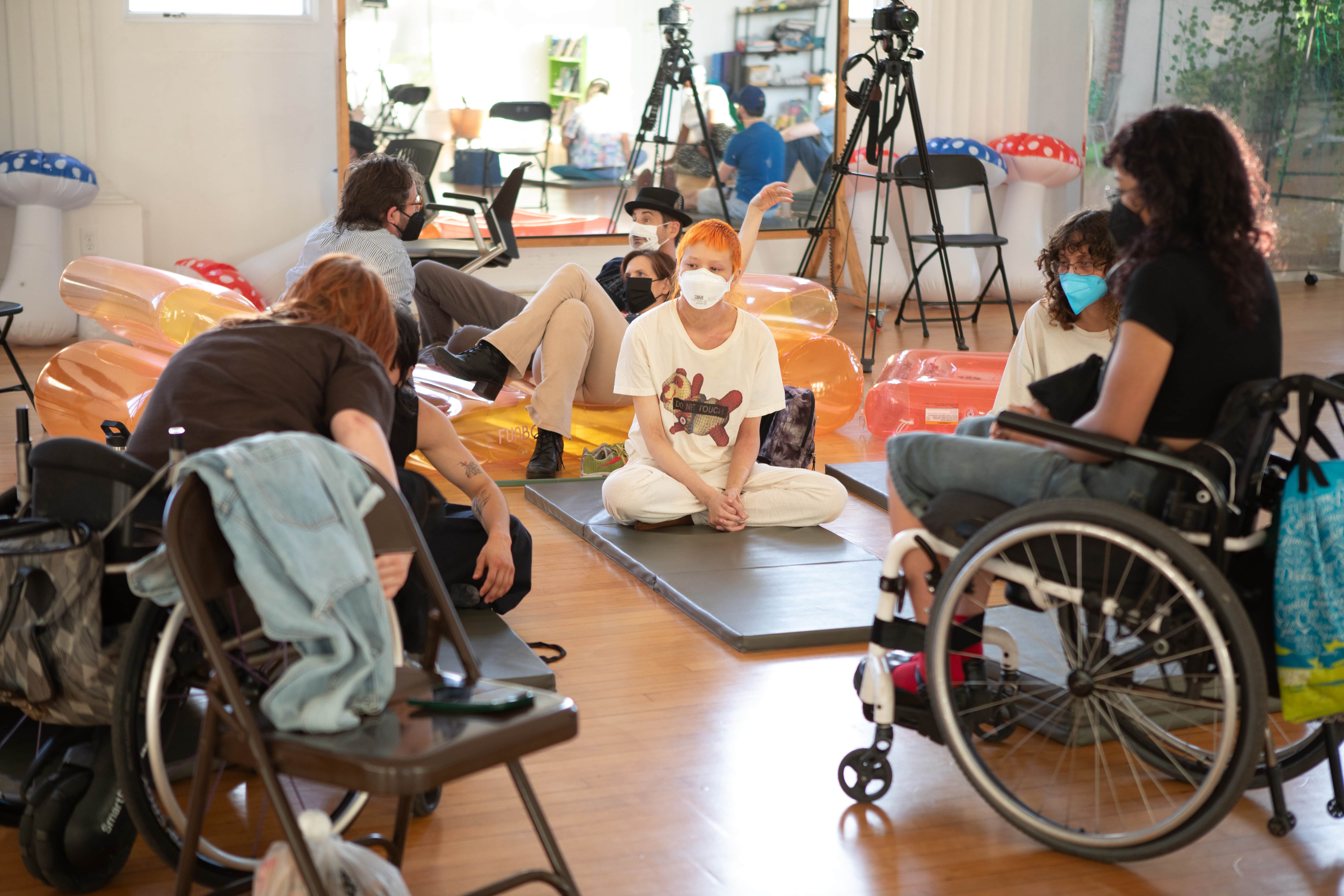 A group of people, some in wheelchairs and some seated on mats on the floor, are gathered in a large, sunlit room. They are all wearing face masks and appear to be in a workshop or class setting, with inflatable objects and camera equipment in the background.