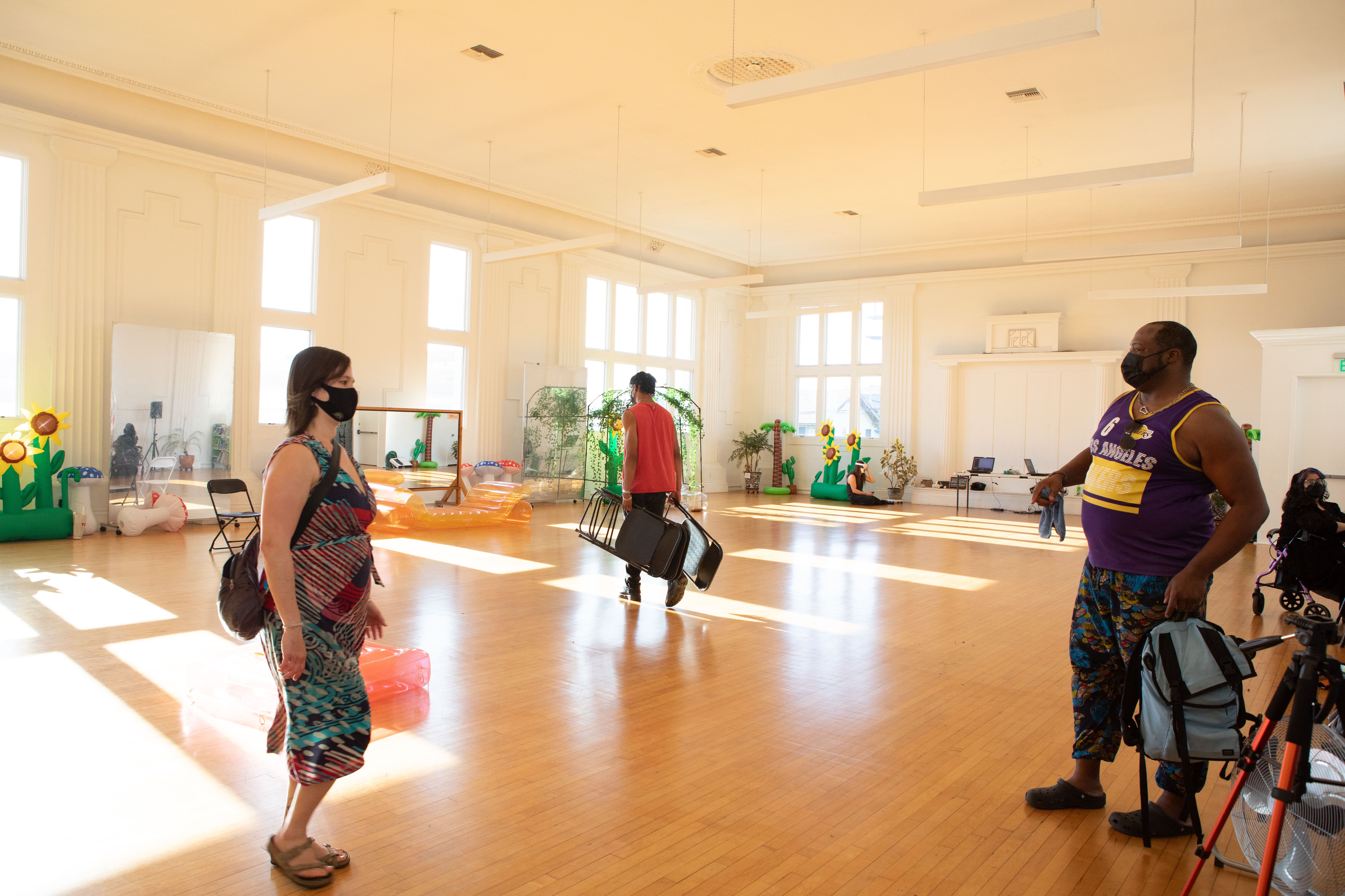 A wide shot of a large, sunlit event hall with polished wooden floors. Three people wearing face masks are visible. A woman in a patterned dress stands in the foreground, a man in a red shirt carries equipment in the middle of the room, and a man in a purple and yellow basketball jersey stands on the right next to a tripod. Inflatable decorations are visible in the background.