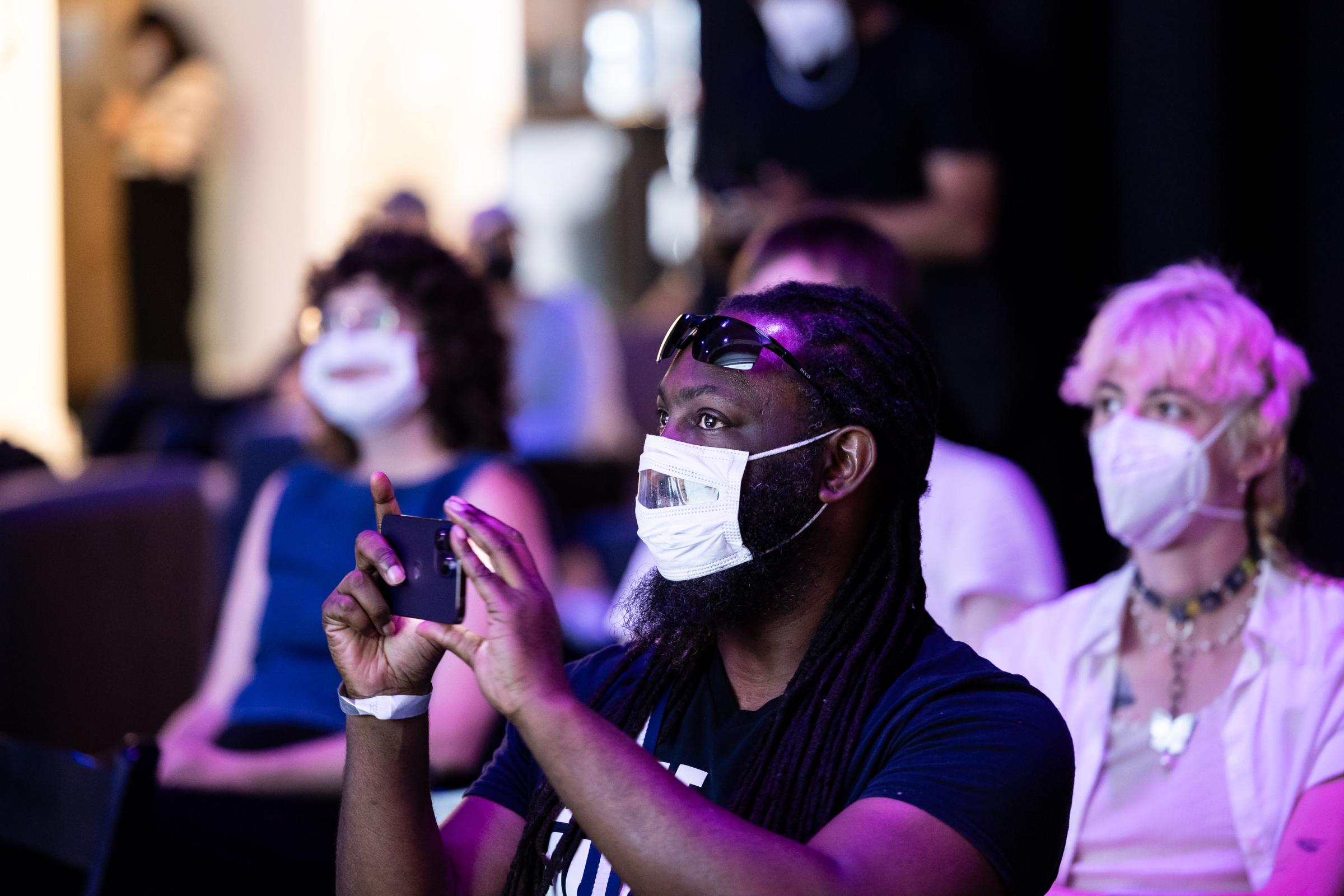Male presenting individual with black hair is taking a video landscape style. Has sunglasses on the top of their face and is wearing a white mask with a clear window in front. Behind is other audience members. 
