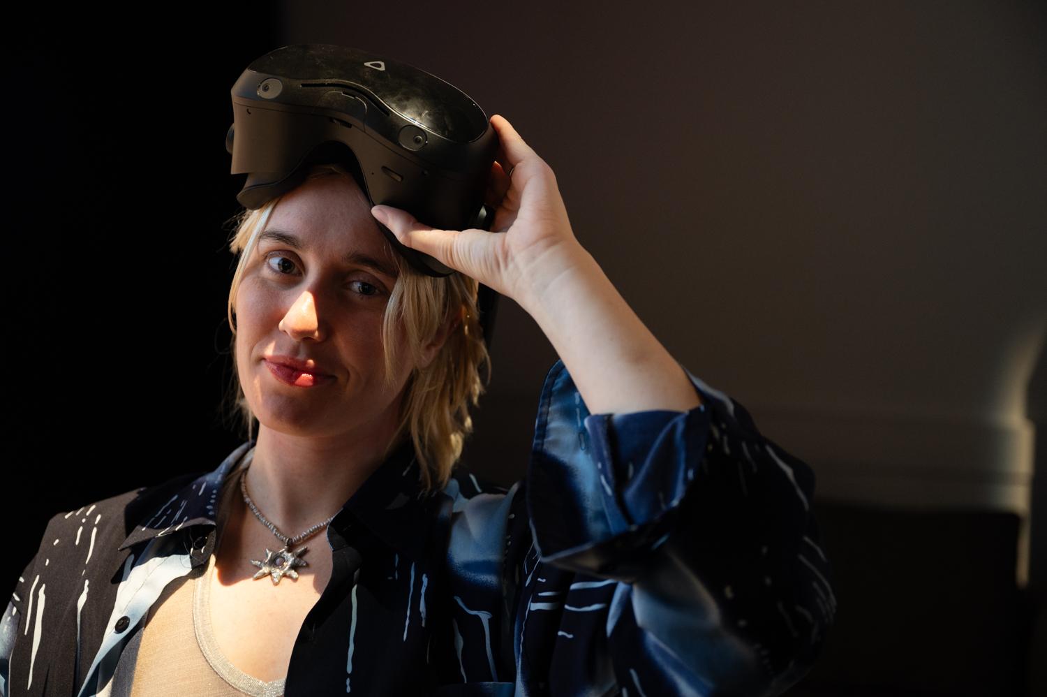 Individual with blonde hair and wearing a leather jacket is lifting their VR headset to look into the camera. 