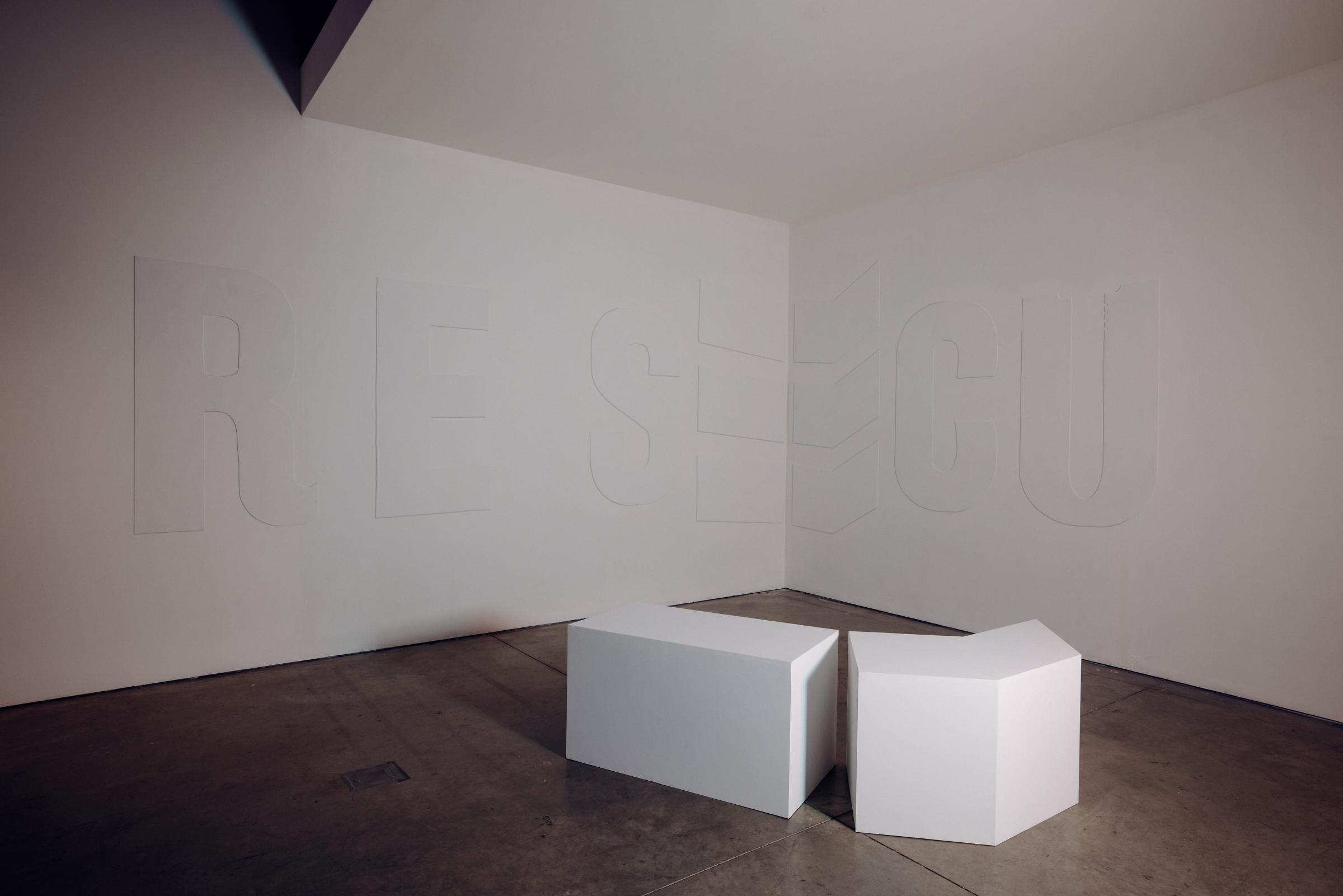 Room with cool light, on the gray colored floor sits two benches in the middle. One is rectangular, the other is a six sided bench with a sharp point. 