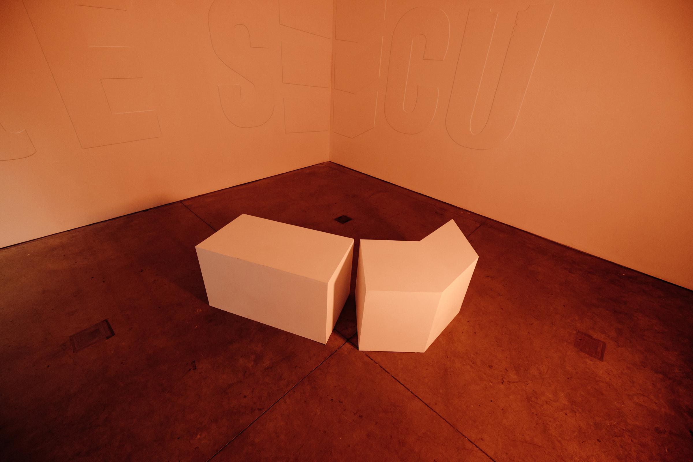 Room with amber glow, on the gray colored floor sits two benches in the middle. One is rectangular, the other is a six sided bench with a sharp point. 