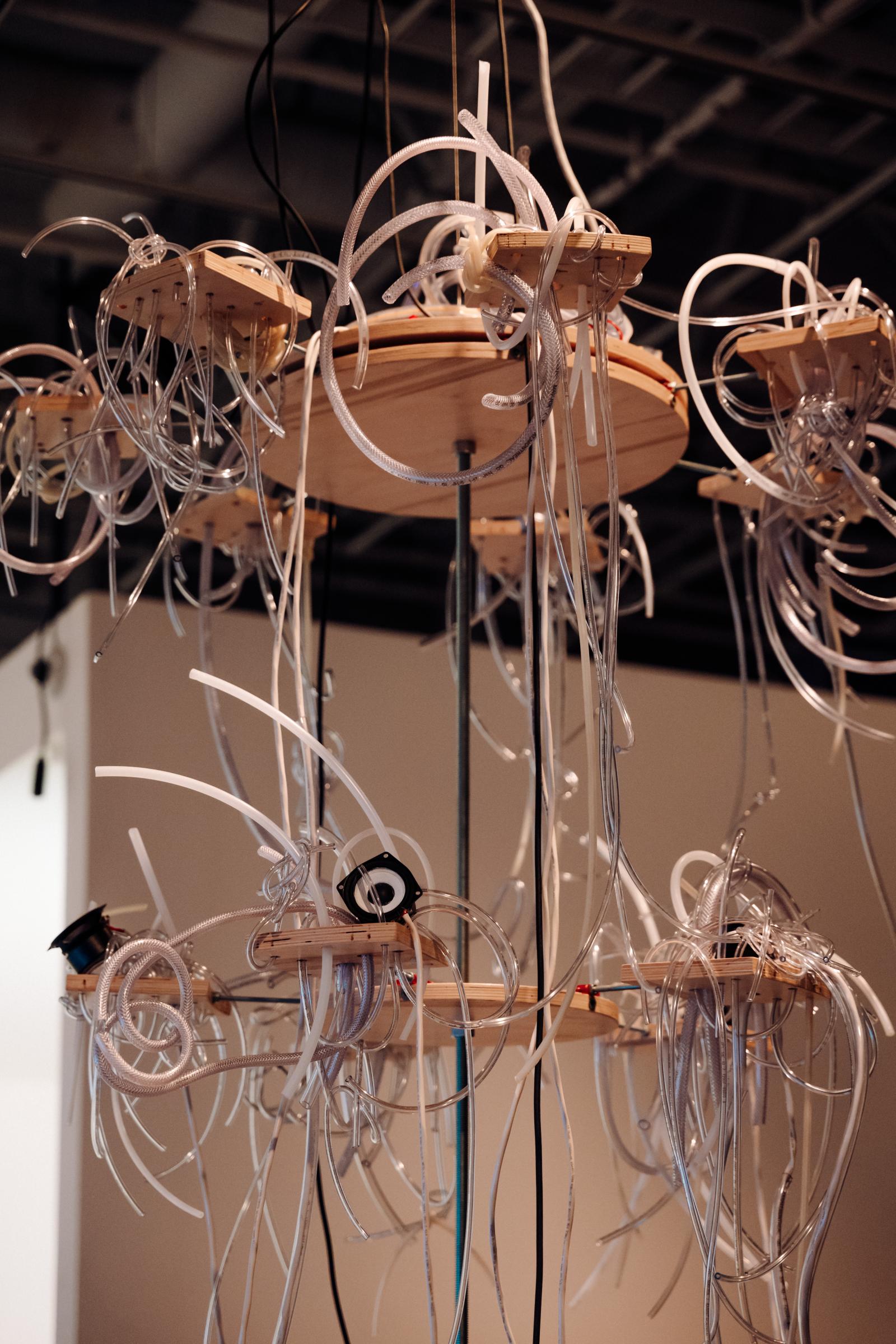 Multi-tiered suspended wooden chandelier with plastic tubes spilling outwards over the sides. There are four speakers and two microphones integrated into the sculpture.