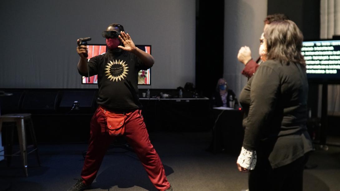 Antoine Hunter, A Black and Indigenous person with dark chocolate skin from his mother,  is wearing a black tshirt, long orange pants and a VR headset. He points one controller upwards in one hand. 
