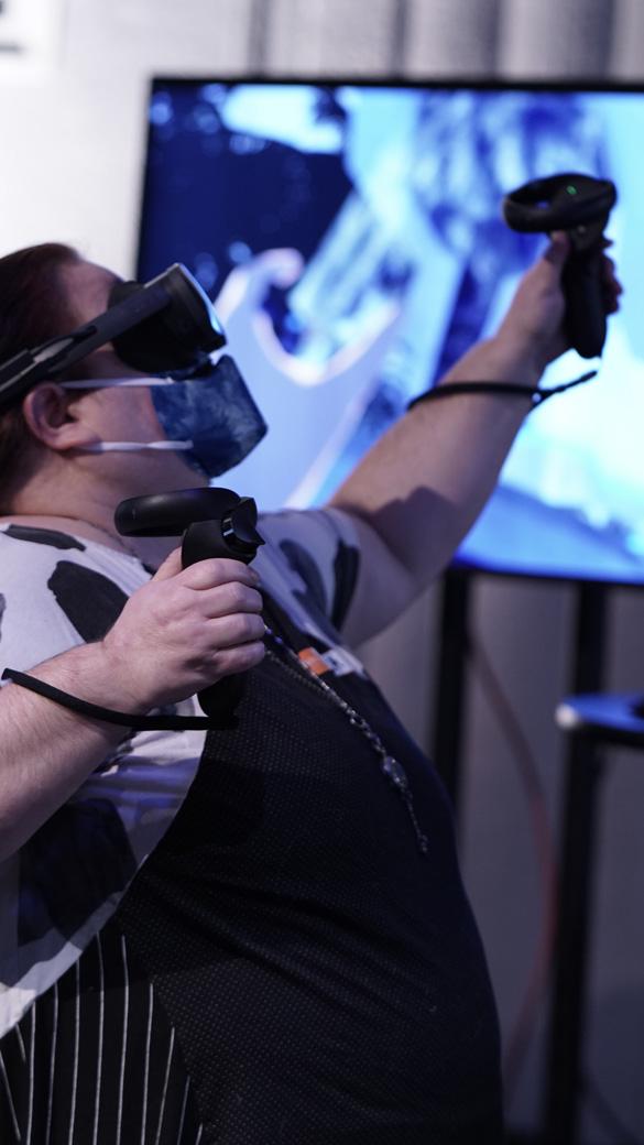 Caucasian fat woman with long auburn hair wearing a black and white shirt with stripes. She is wearing a VR headset and a mask and holding two controllers pointed upwards
