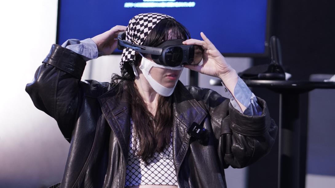 Nat, a white non-binary person with dark brown long hair and step bangs. They are wearing a leather jacket with black and white checkered shirt under and a matching checkered head band. They are adjusting the VR headset they are wearing.