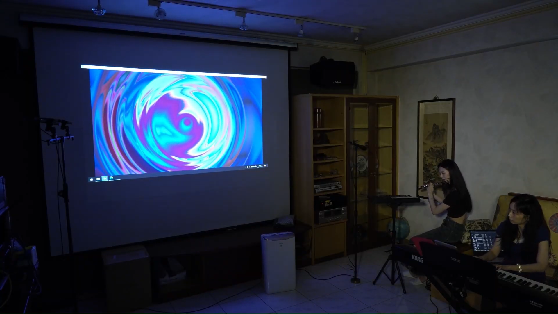A projector showed a sound visualization of concentric circles. Two woman sit in the lower right corner. One plays a flute, the other plays a piano.