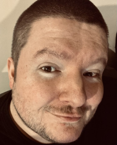 Close-up of the face of a man with short dark hair, trimmed facial hair, and a slight smile, looking directly at the camera under warm lighting. The background is a neutral light colour and slightly blurred.