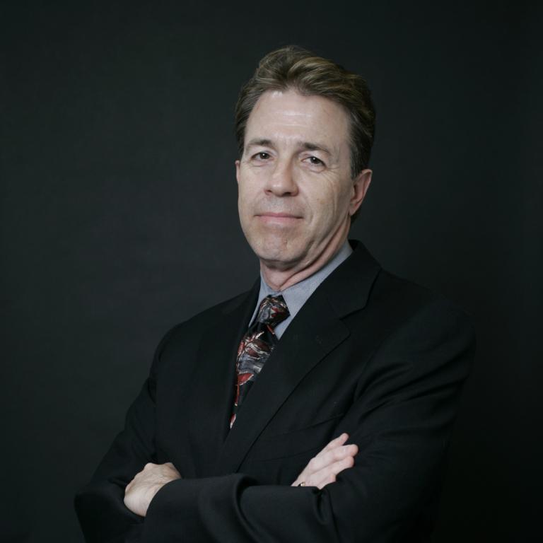 Photograph of Marc Hebert facing the camera, wearing a suit and tie with arms crossed.